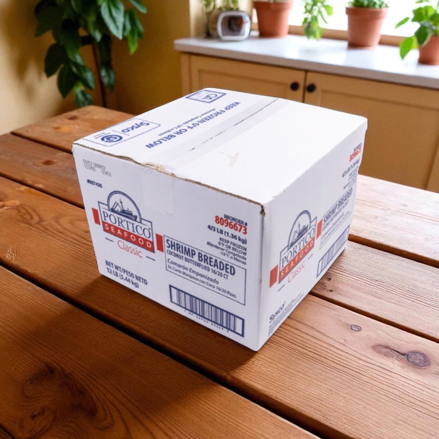 A white Sysco box labeled "Shrimp White Peeled And Deveined Tail-Off 2.5 lb case - 4 Bags" sits on a wooden kitchen table near a window with potted plants.