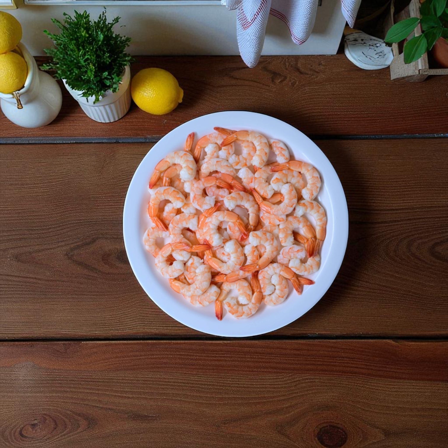 A Sysco Shrimp White Peeled And Deveined Tail-on Cooked 2.5 lb Case (4 Bags) is served on a white plate atop a wooden table, garnished with lemons and surrounded by kitchen items and a potted plant.