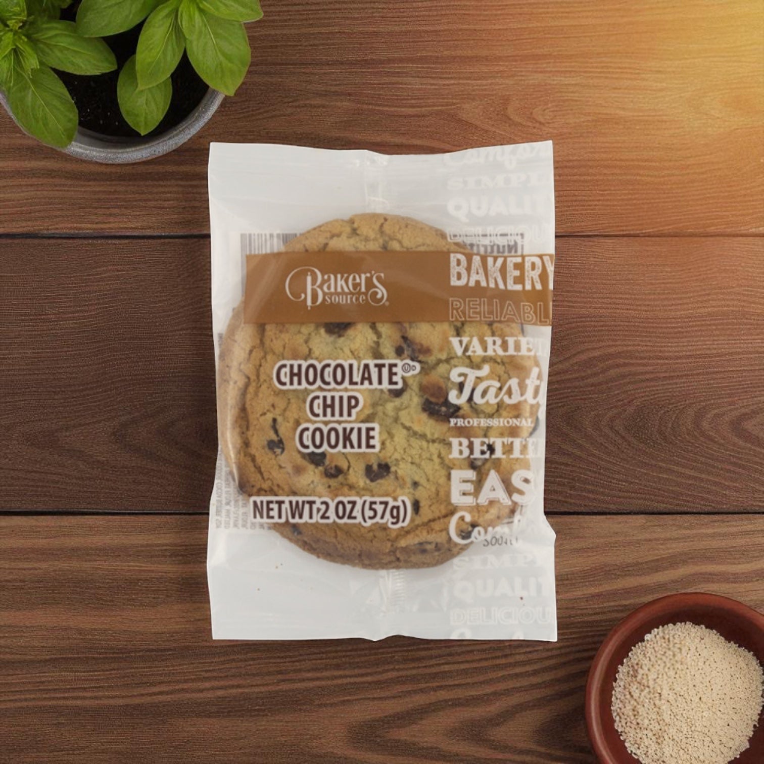 A Baker's Source Classic Chocolate Chip Cookie (2 oz, individually wrapped, 48 count) rests on a wooden table near a plant and brown sugar, highlighting the freshness and convenience of single-serve packaging.
