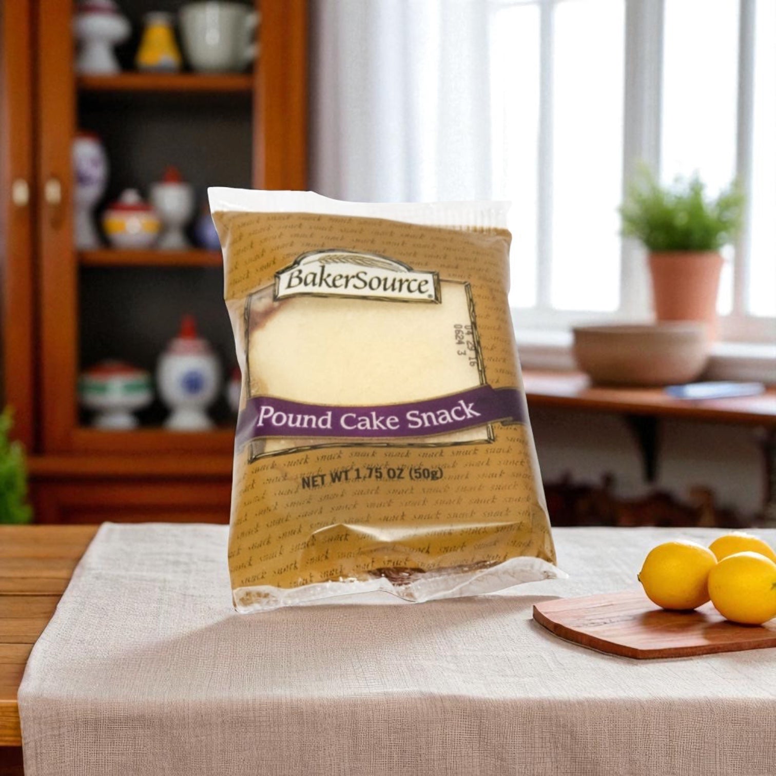 A Baker's Source Classic Cake Pound Individually Wrapped 1.75 oz sits upright on a beige cloth by lemon slices, ideal for grab-and-go, with a cabinet and window in the background. Pack includes 24 cakes.