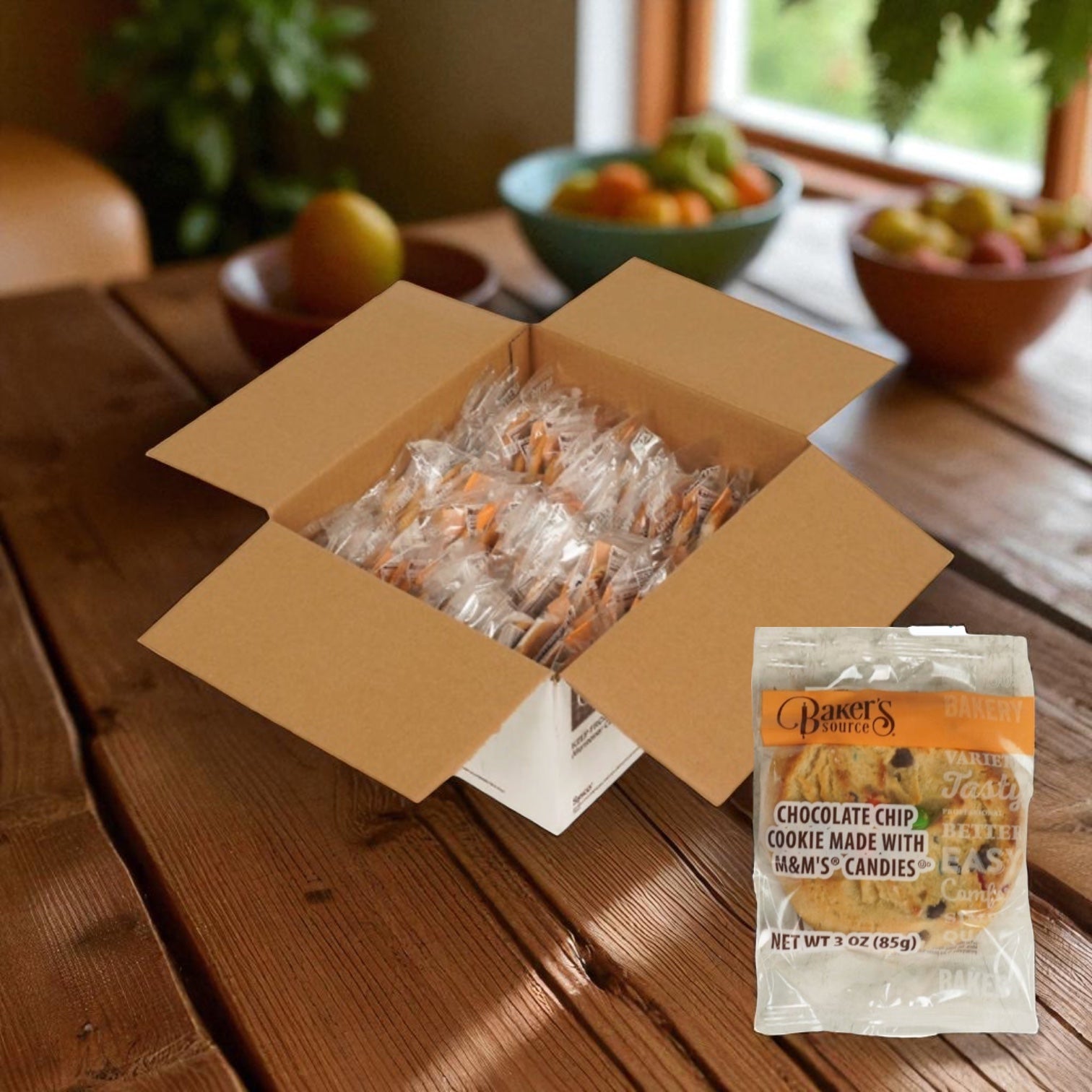 An open box of Baker's Source Classic Cookie Chocolate Chip With Candy, 3 oz individually wrapped (48 count), sits on a wooden table with fruit in the background—perfect for sharing or stocking up with bulk cookies.