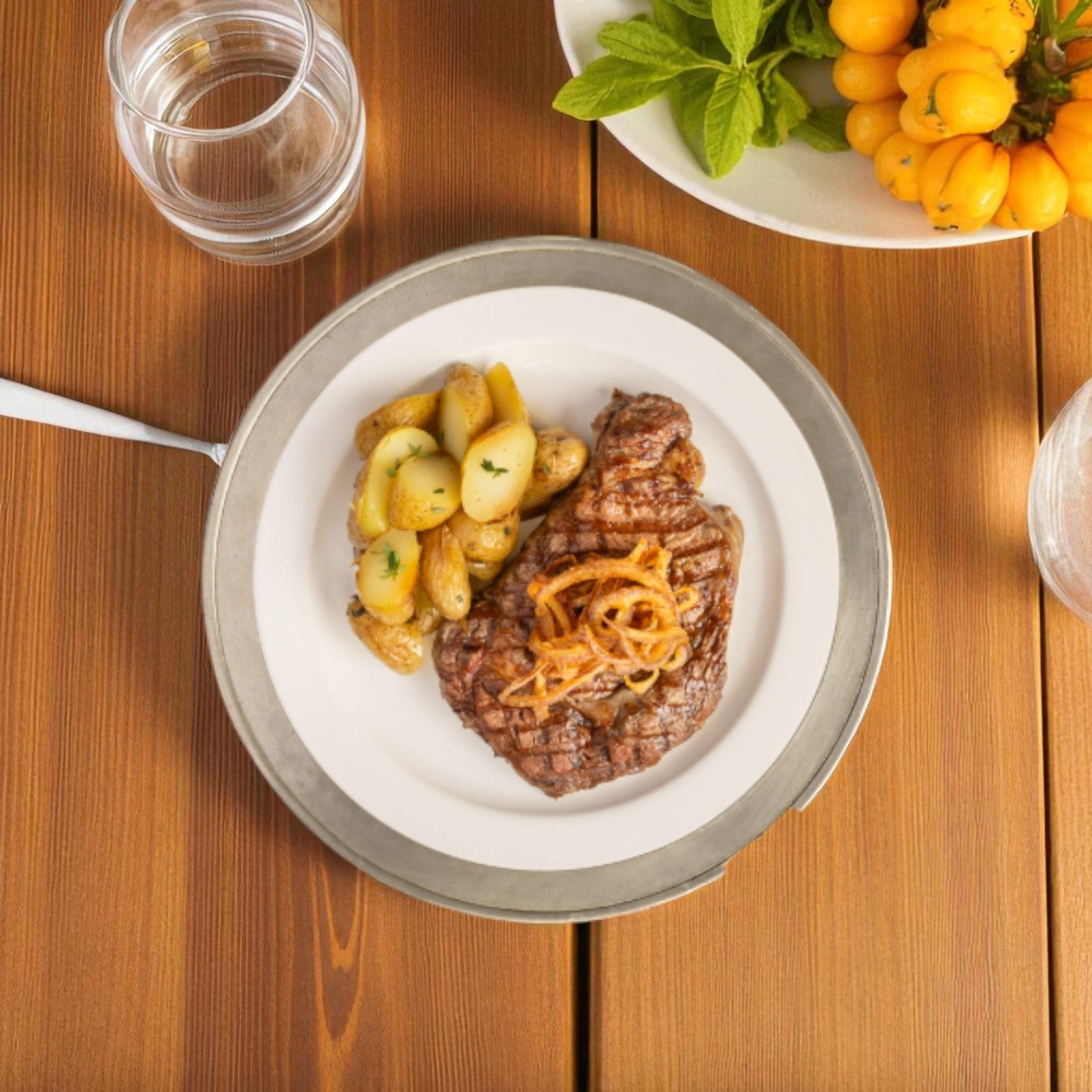A plate with Verde Farms Organic, 100% Grass-Fed Beef Ribeye Steak topped with onions and roasted potatoes sits on a wooden table next to a glass of water and a bowl of yellow fruits with herbs.