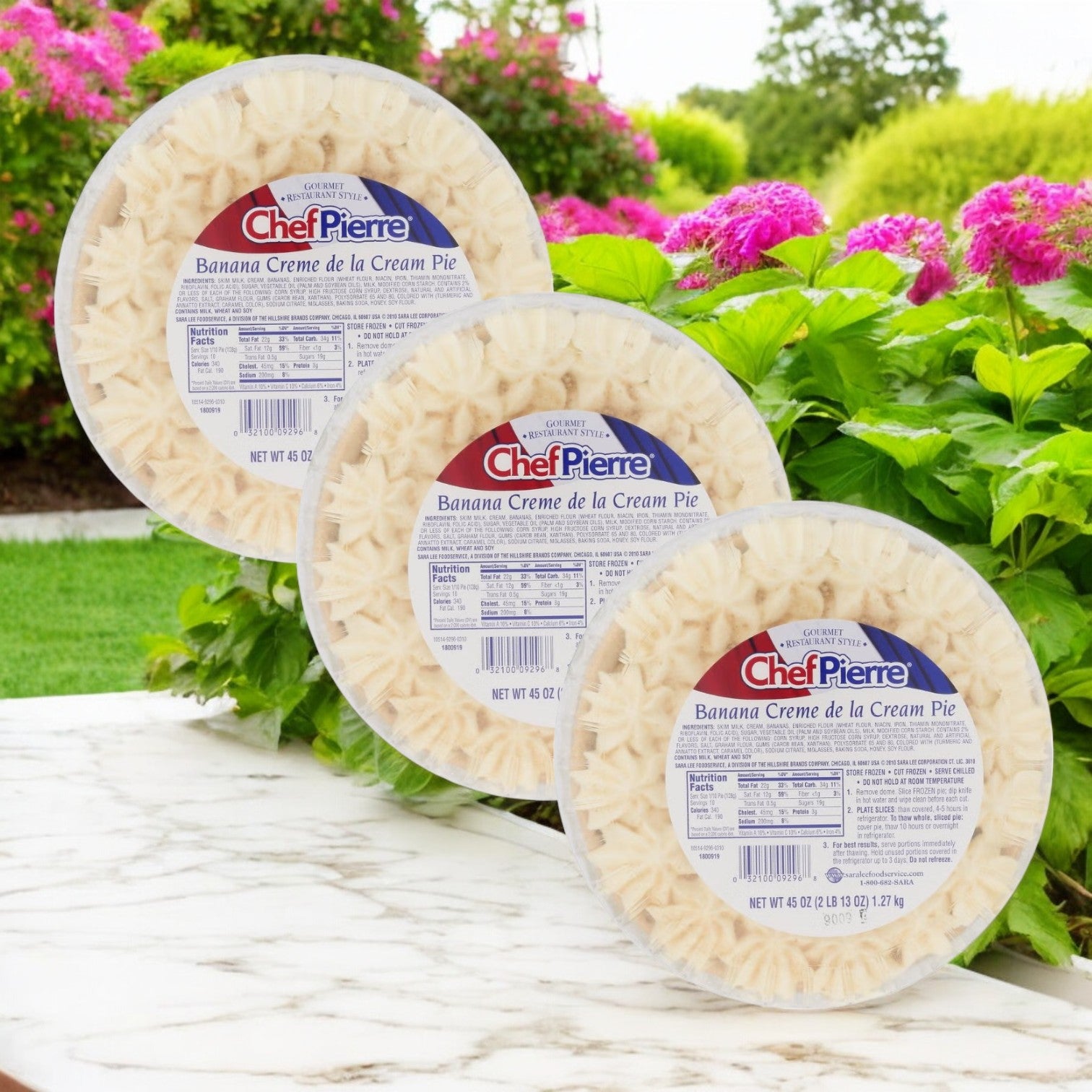 Three Chef Pierre Banana Crème de la Cream Pies, each 10 inches and boasting a rich banana filling, are displayed outdoors among flowers and greenery. This delightful frozen dessert from Chef Pierre is perfect for any occasion.