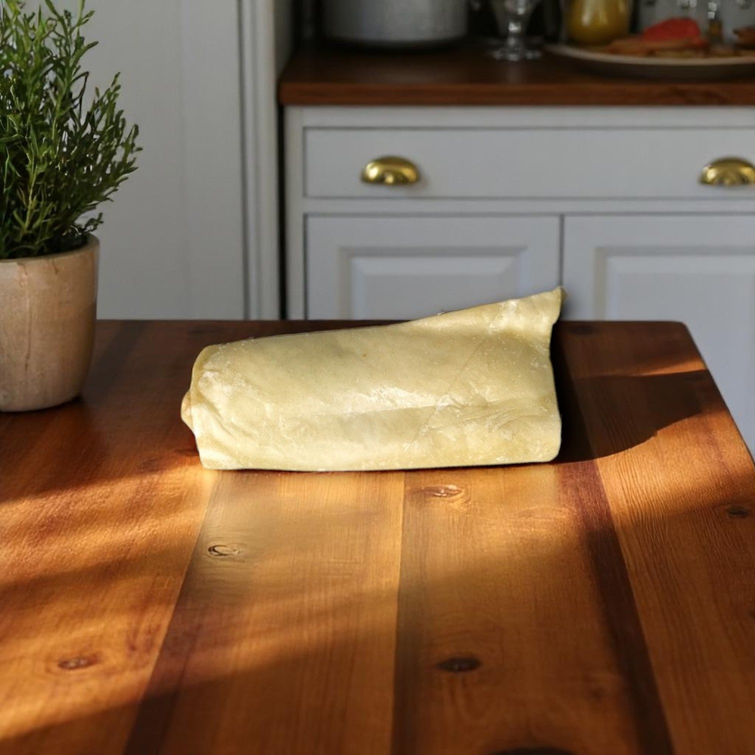 An individually wrapped Iltaco Beef & Bean Burrito – 5 oz from The Original Pizza Puff sits on a wooden kitchen table, ready to enjoy, with a potted plant and white cabinets in the background.