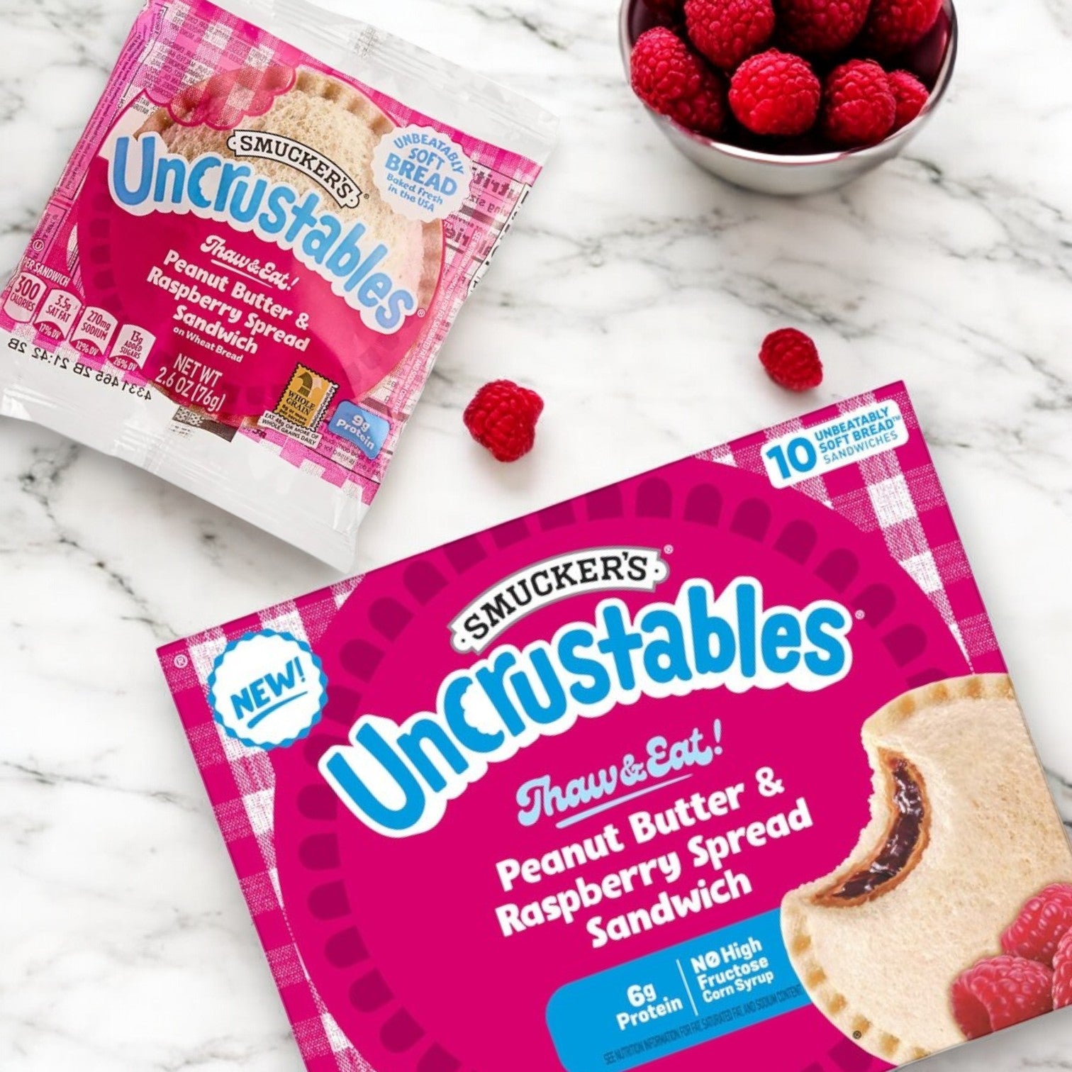 An Uncrustable Smuckers Peanut Butter & Raspberry Spread Sandwich sits next to a 10-pack box and fresh raspberries, showcasing the convenient Smucker's Peanut Butter & Raspberry Spread Sandwich – 10 Pack (2 oz Each).