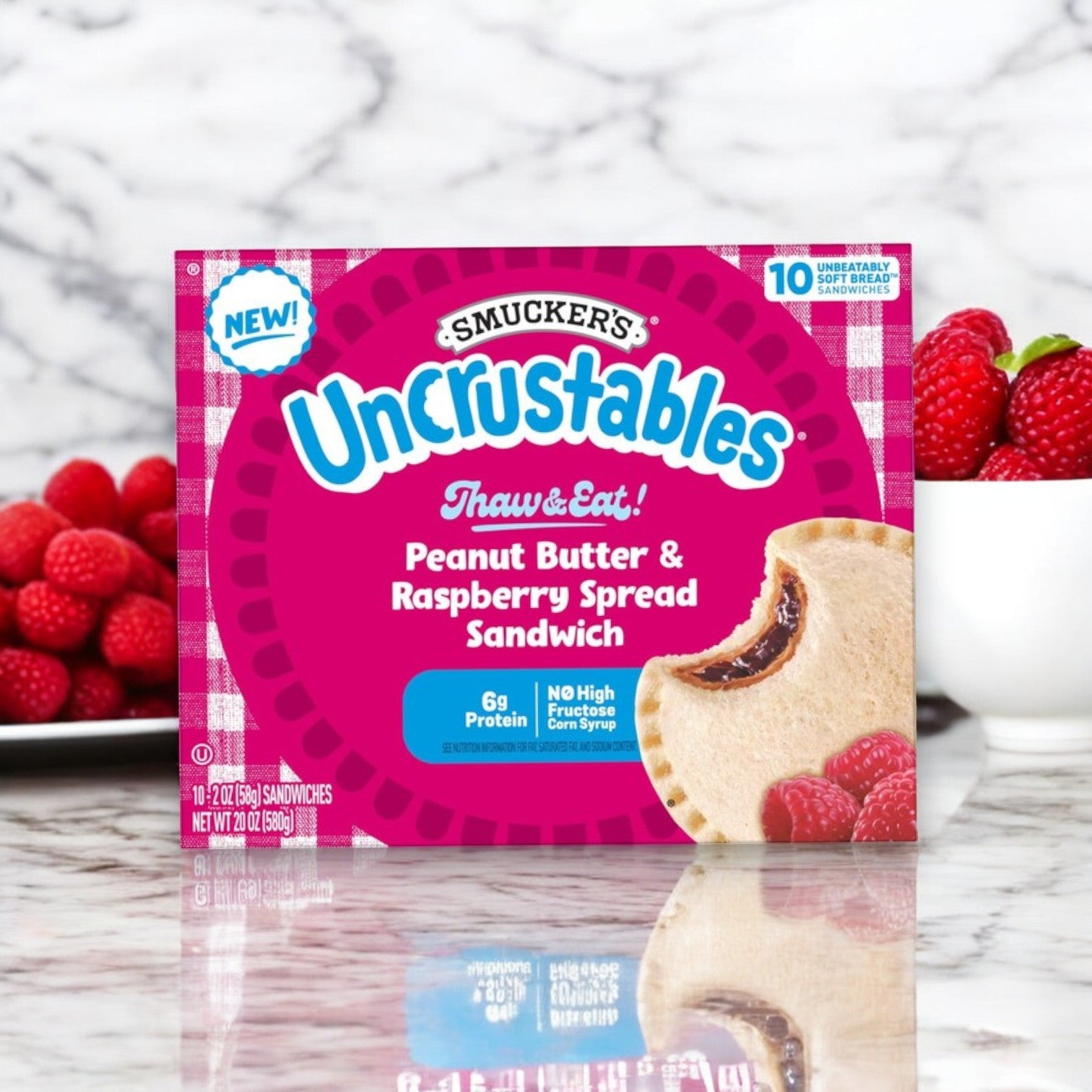 A box of Uncrustable Smuckers Peanut Butter & Raspberry Spread Sandwiches – 10 Pack (2 oz each) sits on a table, offering a convenient snack, with fresh raspberries and a white bowl in the background.