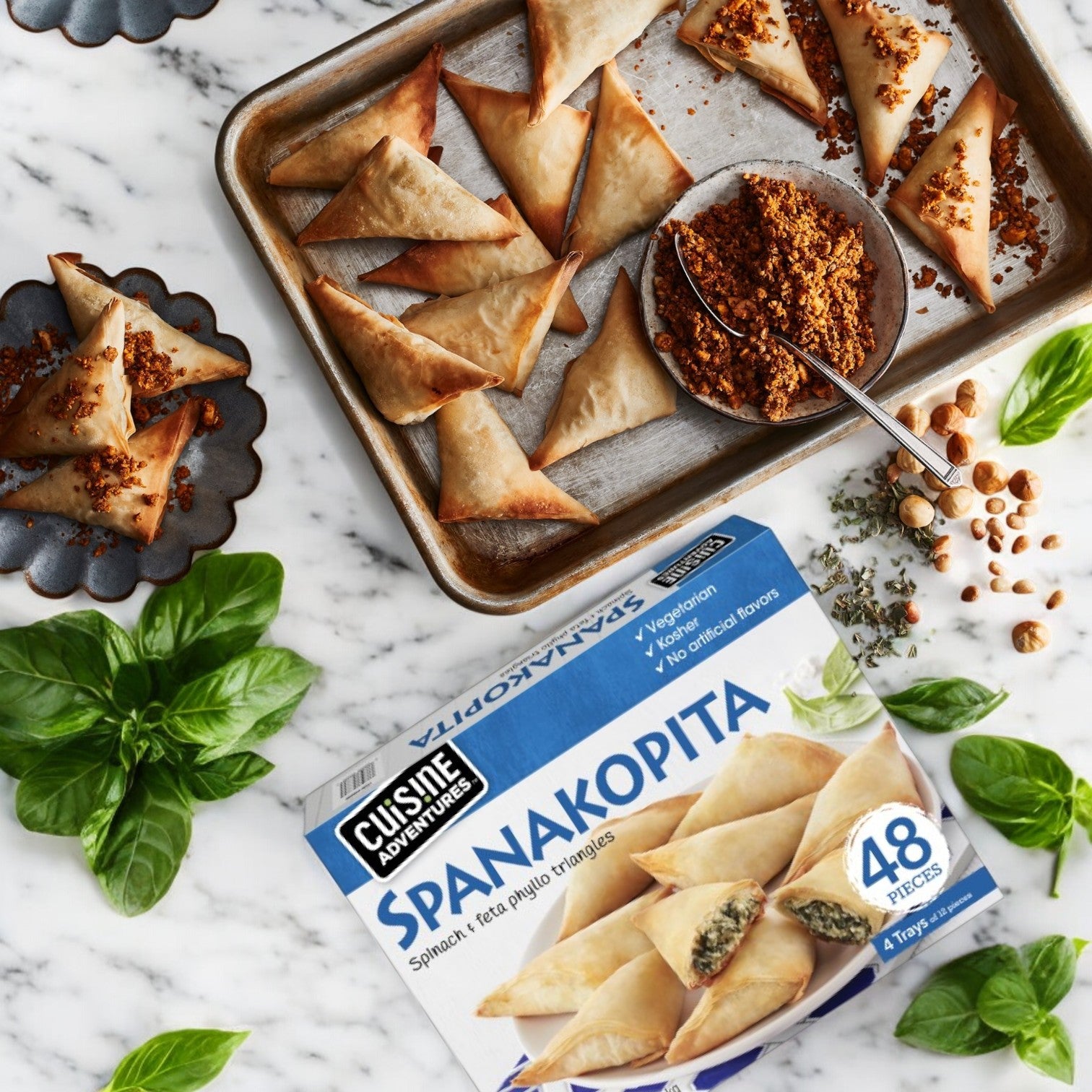 A tray of Cuisine Adventures Spanakopita Spinach & Feta Phyllo Triangles sits beside a box labeled "Cuisine Adventure," offering 48 pieces. Crumbled topping, basil leaves, and chickpeas are scattered across the marble surface.