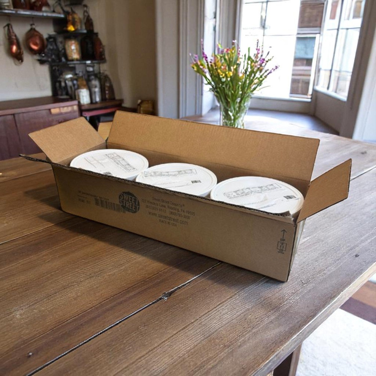 An open box with three white tubs of Sweet Street Scoopable Apple Compote (102 oz each) sits on a kitchen table with sunlight and flowers in the background—ideal as a dessert topping.