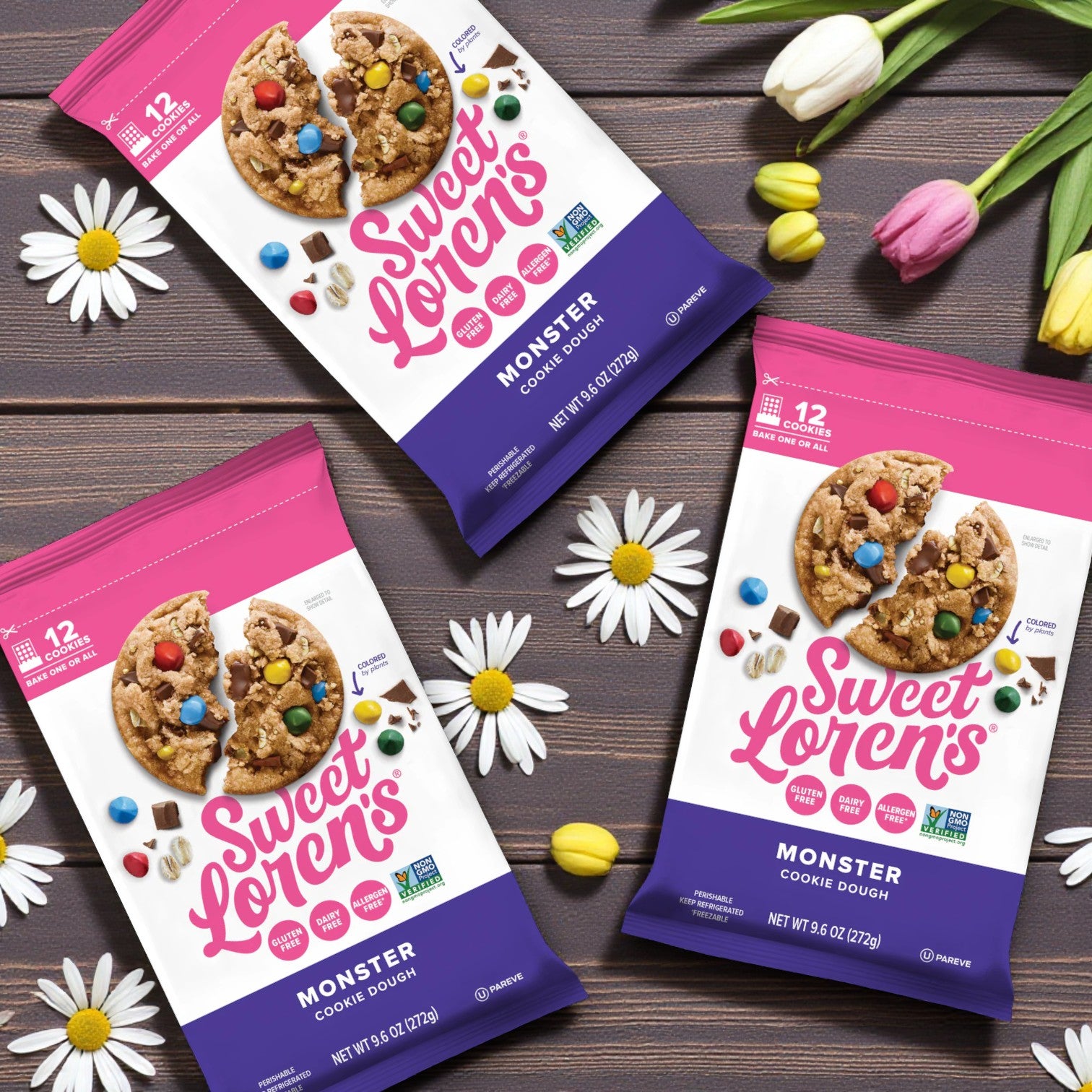 Three packs of Sweet Loren’s Monster Cookie Dough – 9.6 oz each, are displayed on a wooden surface with scattered daisies and green leaves.
