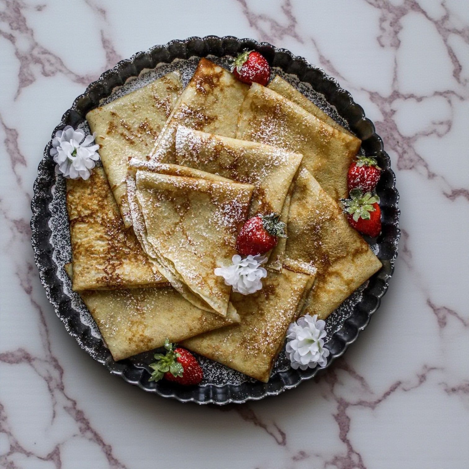 Sysco Premium Plain Crepes, 6", from Sysco are displayed folded on a round metal tray, dusted with powdered sugar and garnished with strawberries and white flowers, set on a marble surface. Pack includes 5 layers, 20 count.