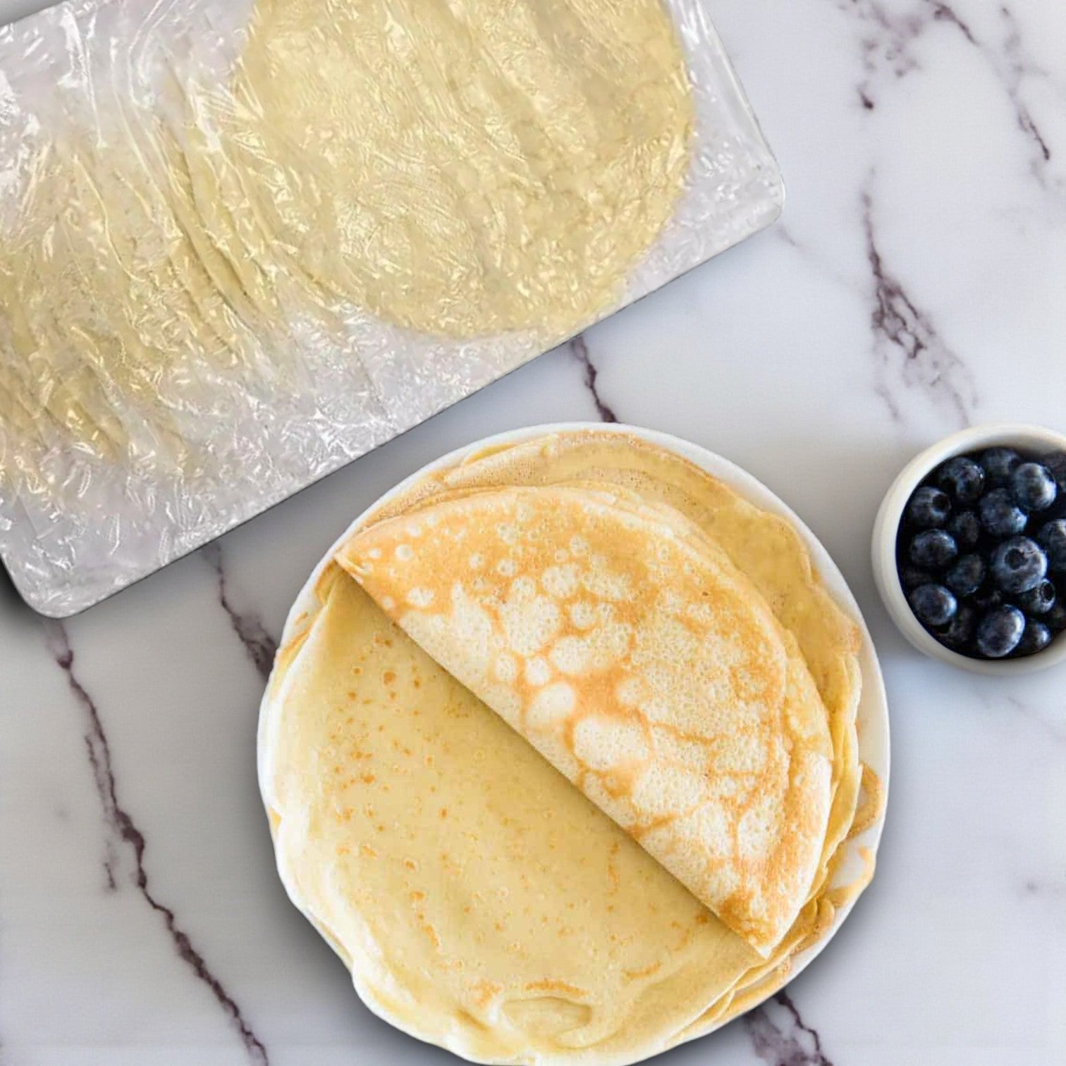A plate of plain crepes, one folded, sits beside a package of Sysco Premium Plain Crepes, 6" – 5 Layers / 20 Count, and a small bowl of blueberries on a marble surface.
