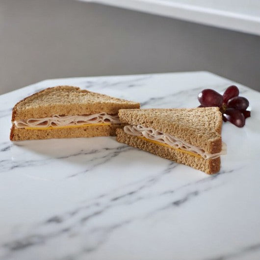 An E.A. Sween Oven Roasted Turkey & Cheese Sandwich Wedge sits halved on whole wheat bread beside red grapes on a white plate, with a 30-day shelf life post-freezer.