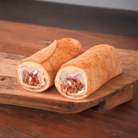 Two halves of an E.A. Sween Turkey Club Wrap—stuffed with bacon, ham, cheddar cheese, and ranch dressing—rest on a wooden cutting board. Ships frozen in an 8-count case; 30-day shelf life in the cooler once thawed.