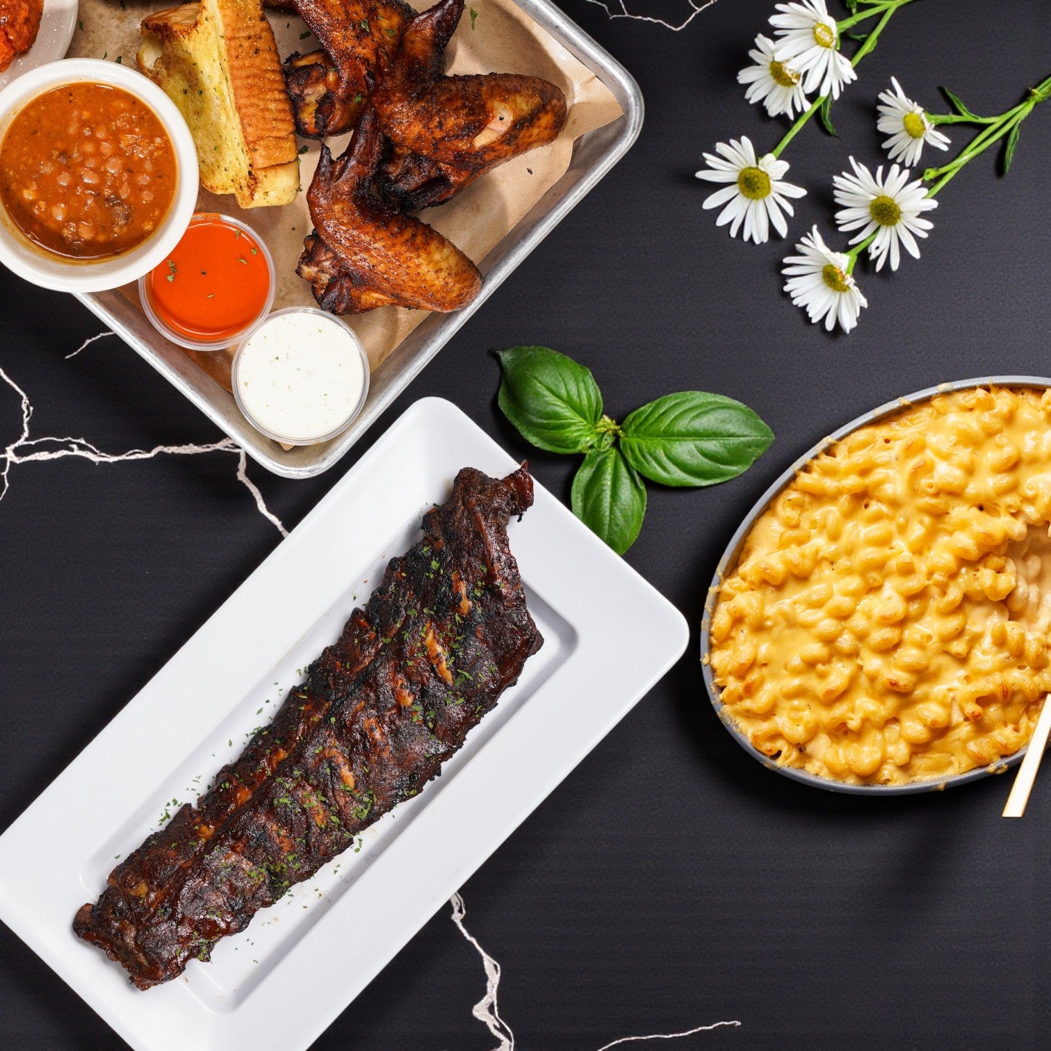 Savor the TJ Ribs - Full Single Dinner by TJ Ribs, featuring barbecued ribs, chicken wings, baked beans, dipping sauces, and macaroni & cheese—served together for the perfect BBQ meal.