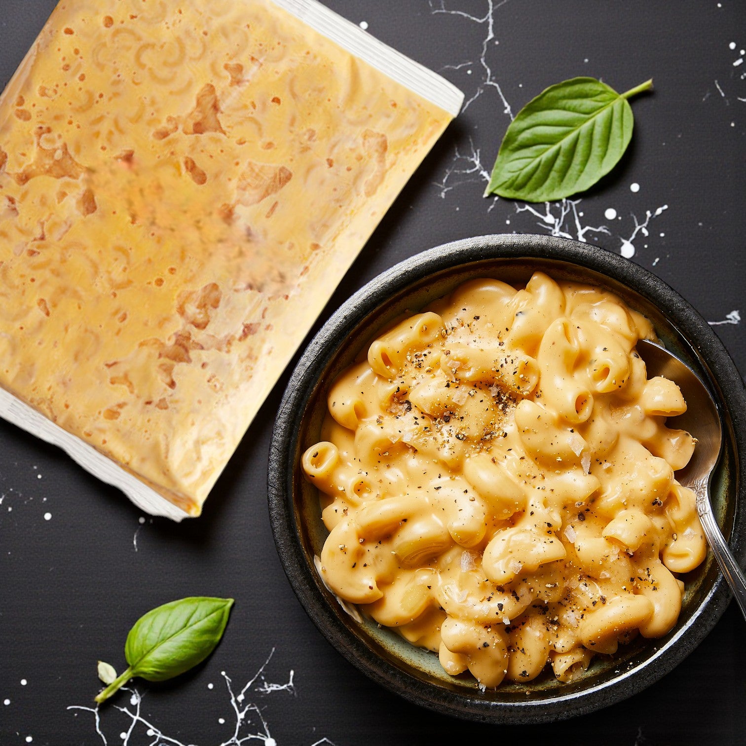 A bowl of TJ Ribs Mac & Cheese – 6 oz by TJ Ribs sits beside a sealed cheese sauce packet, garnished with basil leaves on a black surface—the perfect comforting individual side dish.