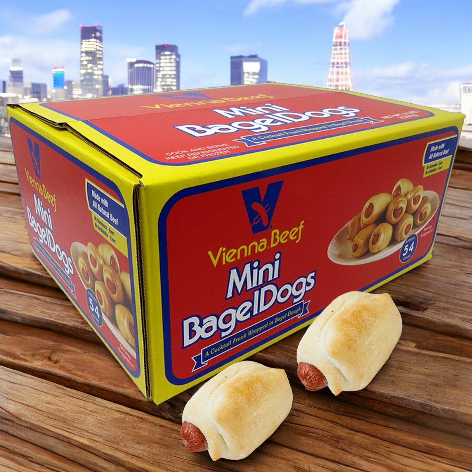 A box of Vienna Beef Mini Bagel Dogs (54 Mini Bagel Dogs) by Vienna Beef rests on a wooden surface, with two tasty snacks in front and city buildings visible in the background.