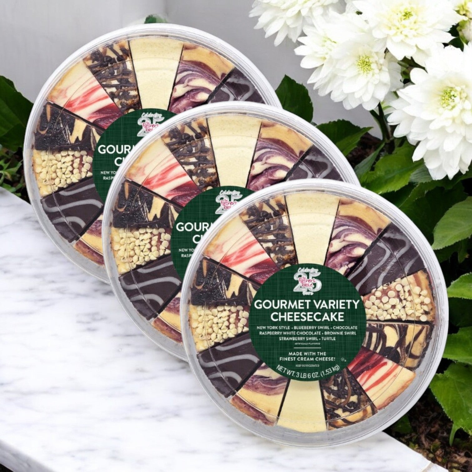 Three packs of The Father’s Table Variety Cheesecake – 10 Inch, 14-Cut, crafted with premium ingredients and various flavors, are elegantly displayed on a marble surface beside white flowers.