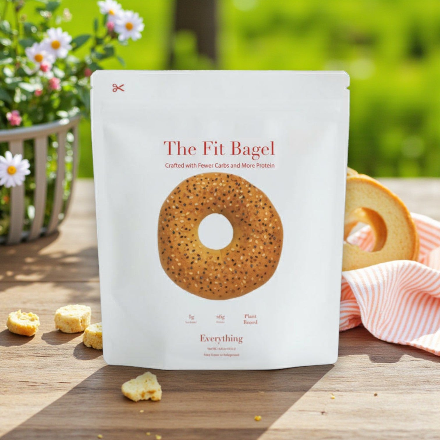 A package of The Fit Bagel – Everything Bagels (4 bagels, The Fit Bagel brand) stands on an outdoor table with a bagel, classic seasoning crumbs, a striped cloth, and flowers in the background.