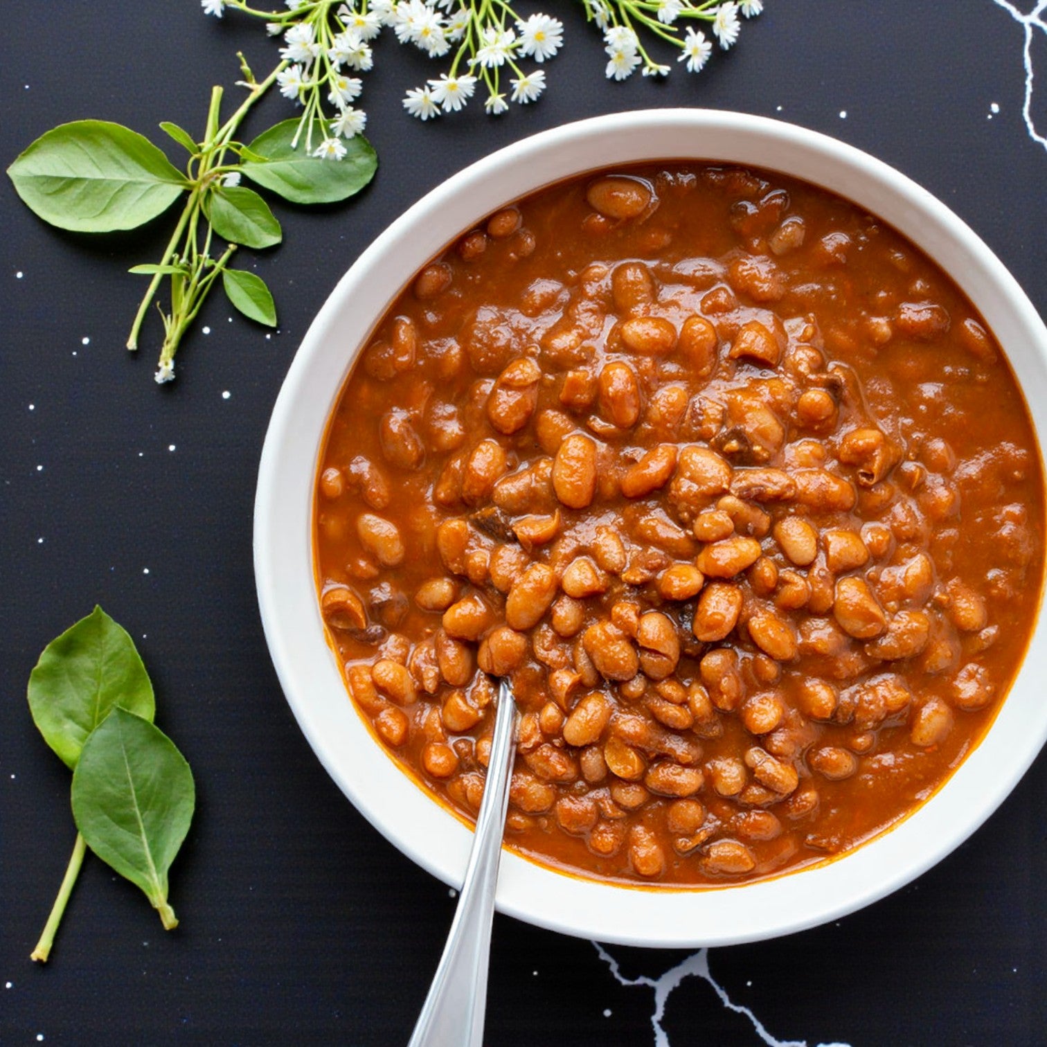 A 6 oz bowl of TJ Ribs Baked Beans by TJ Ribs, served with a spoon on a black surface adorned with green leaves and white flowers.