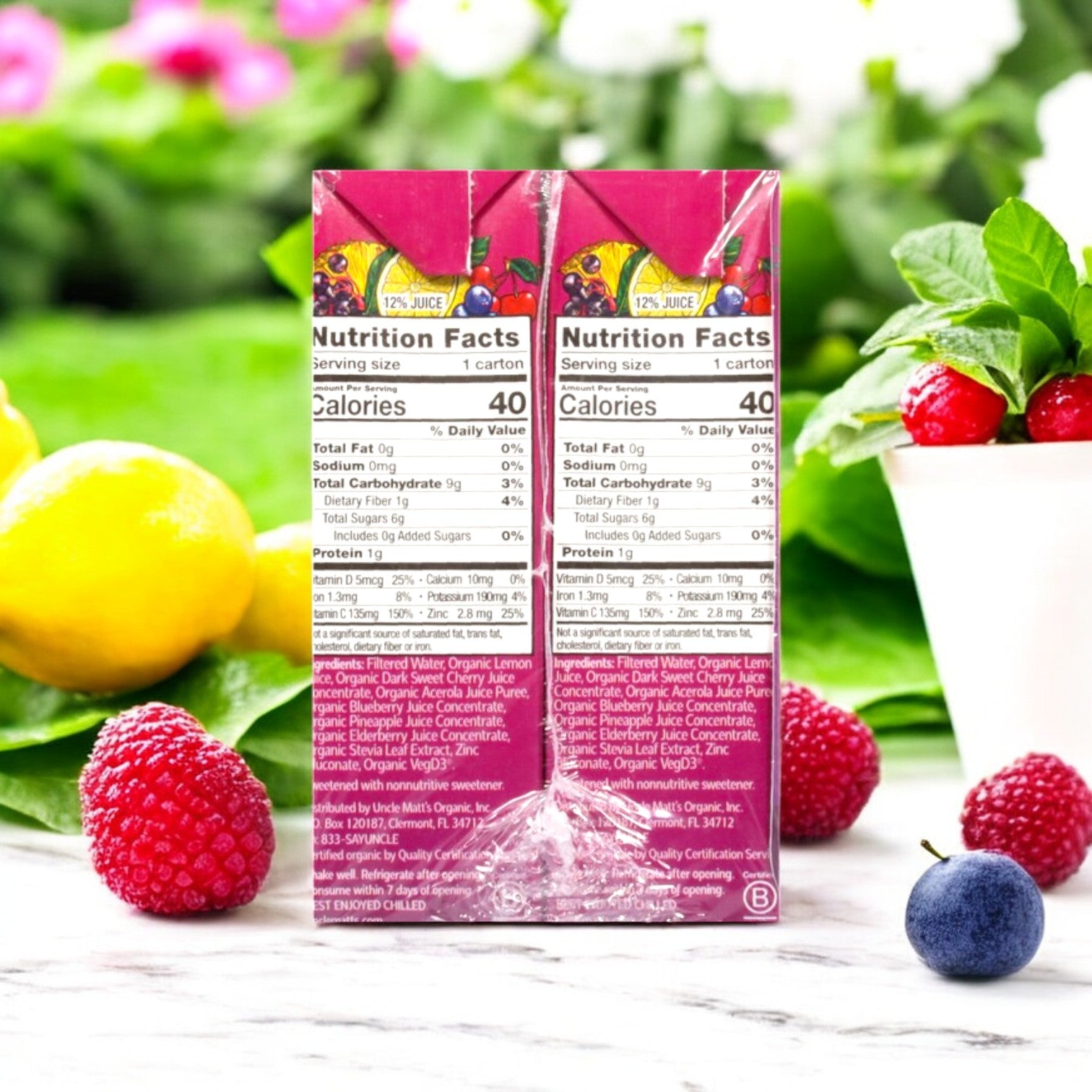 Two Uncle Matt's Organic Superfruit Punch juice boxes displaying "No Sugar Added" are surrounded by fresh raspberries, a blueberry, lemons, and greenery. This organic drink bursts with antioxidants and vitamins, creating a vibrant and nutritious scene.