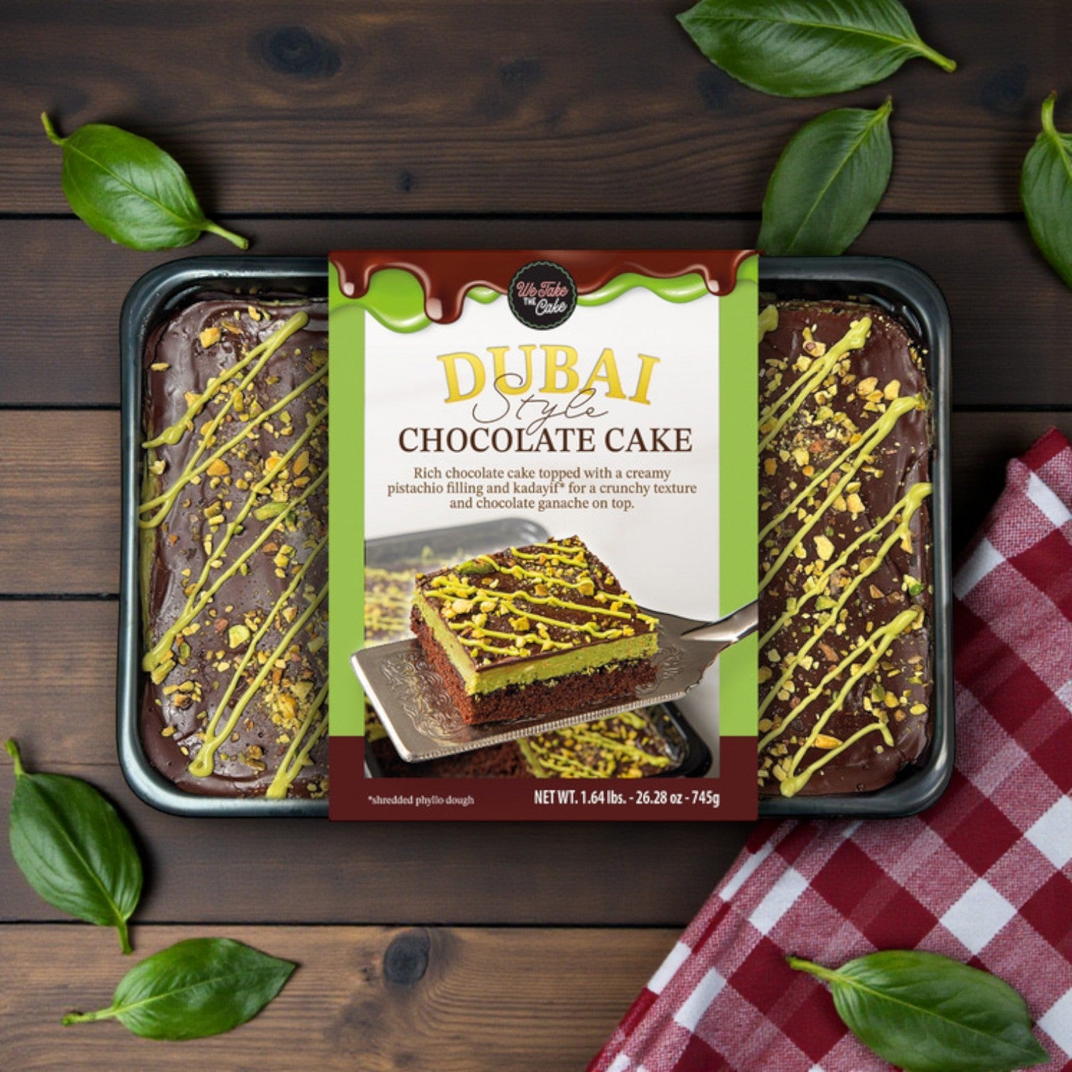 We Take The Cake – Dubai-Style Chocolate Cake, 26.28 oz by We Take The Cake, comes tray-packaged and topped with pistachios and chocolate drizzle for a premium dessert experience. Displayed on wood with green leaves for added elegance.