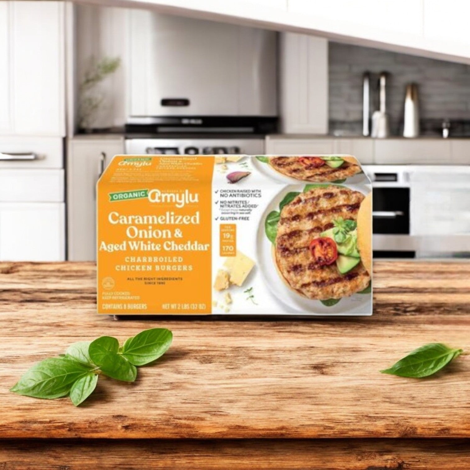 A box of Amylu’s 4-Pack Combo – Premium Chicken & Sausage Variety, featuring antibiotic-free chicken, sits on a wooden kitchen counter beside basil leaves, with kitchen appliances in the background.