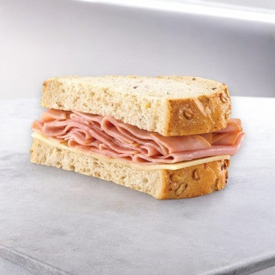 An E.A. Sween Ham & Swiss sandwich on rustic oat-topped wheat bread is displayed on a light gray surface. Ships frozen, 1 case (8 count), and has a 30-day shelf life in the cooler once thawed.