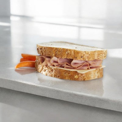 An E.A. Sween Ham & Swiss sandwich on rustic oat-topped wheat bread sits next to sliced tomato. Sold frozen, 1 case (8 count), and lasts 30 days in the cooler once thawed.