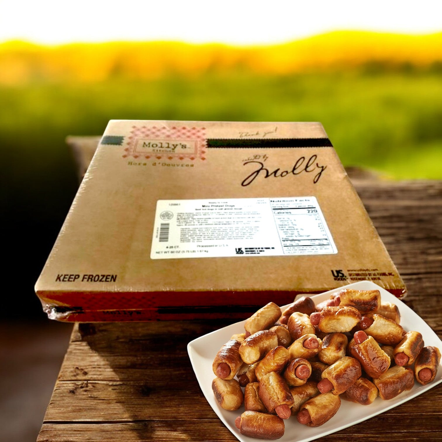 A box of Molly's Kitchen Pretzel Dogs (100 Count, 0.6 oz each, parbaked) rests on a wooden surface beside a white plate with cooked pretzel dogs; the setting features an outdoor backdrop with greenery.