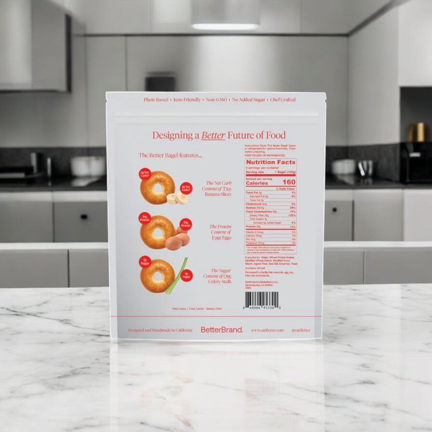 A white BetterBrand food package of The Classic Better Bagel (6 bags, 24 bagels) highlights High Protein and Low Carb features as it stands on a kitchen counter with a blurred modern kitchen in the background.