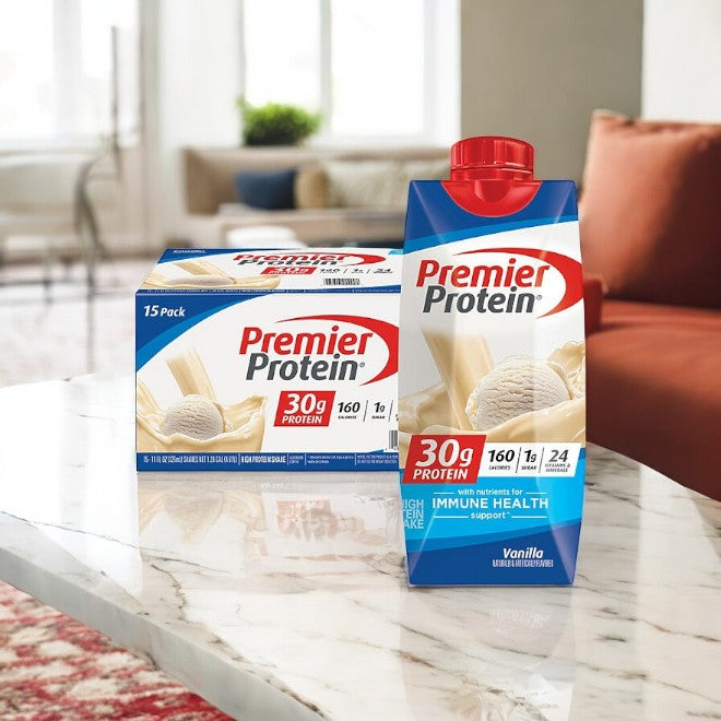 A Premier Protein 30g High Protein Shake, 11 oz, and a 15-pack case are displayed on a marble table in a cozy living room, ideal for post-workout recovery.