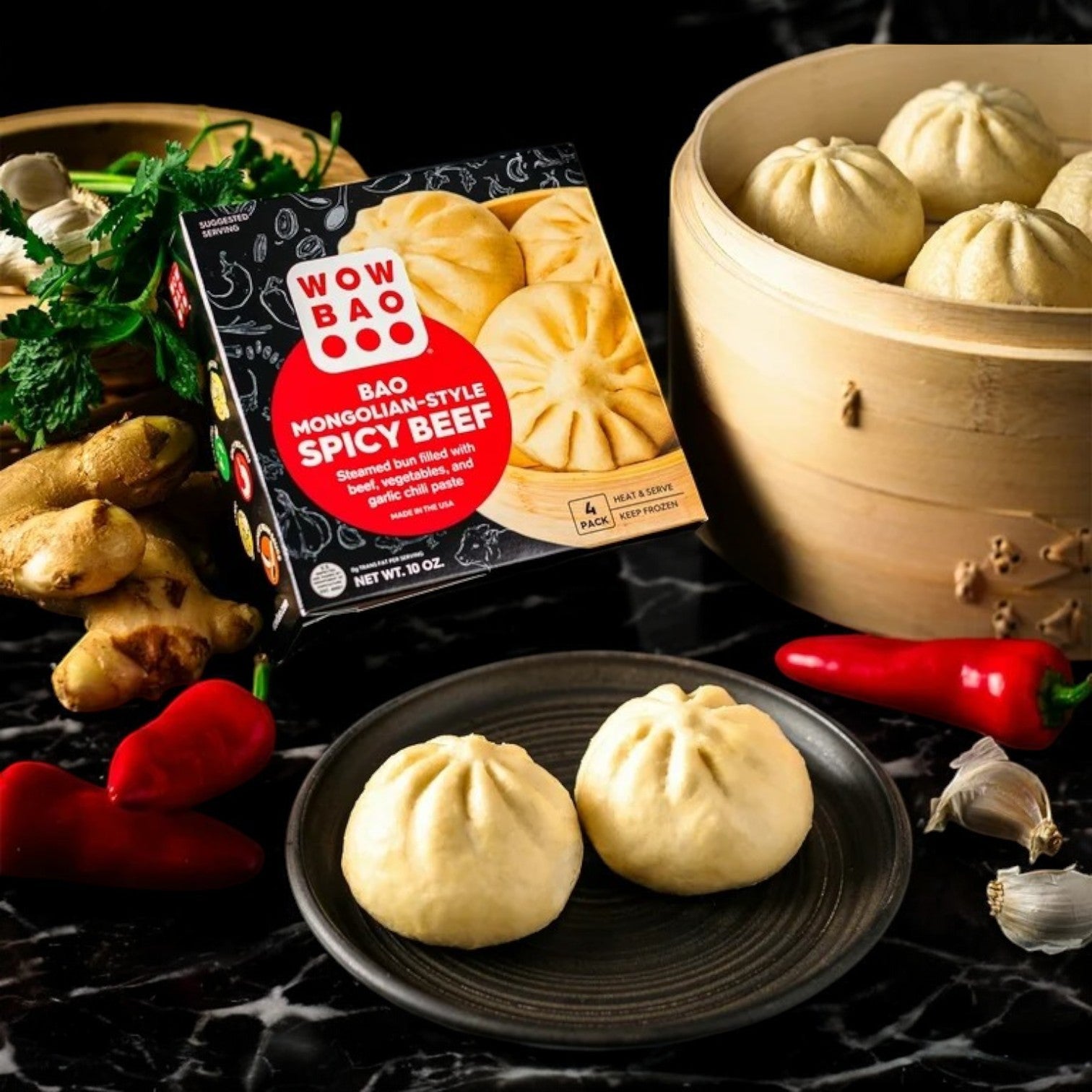 A box of Wow Bao Mongolian-Style Spicy Beef, Bao-1 Count by Wow Bao sits beside a plate and steamer of savory buns, showcasing ginger, garlic, cilantro, and red chili peppers on a black marble surface.