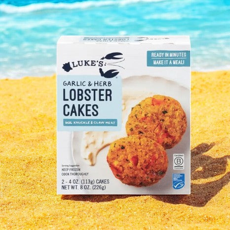 A box of Luke's Organic Lobster Cakes, Garlic & Herb-1 Count by Luke's is displayed on a yellow background, showing two lobster cakes and a serving suggestion with sauce on the packaging.