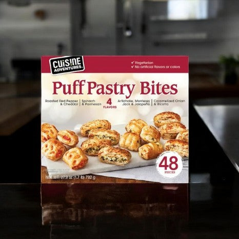 On the counter sits a Cuisine Adventure Puff Pastry Bites Variety Pack, featuring a gourmet selection. With 48 vegetarian pieces in four distinct flavors, it offers indulgence free from artificial flavors or colors.