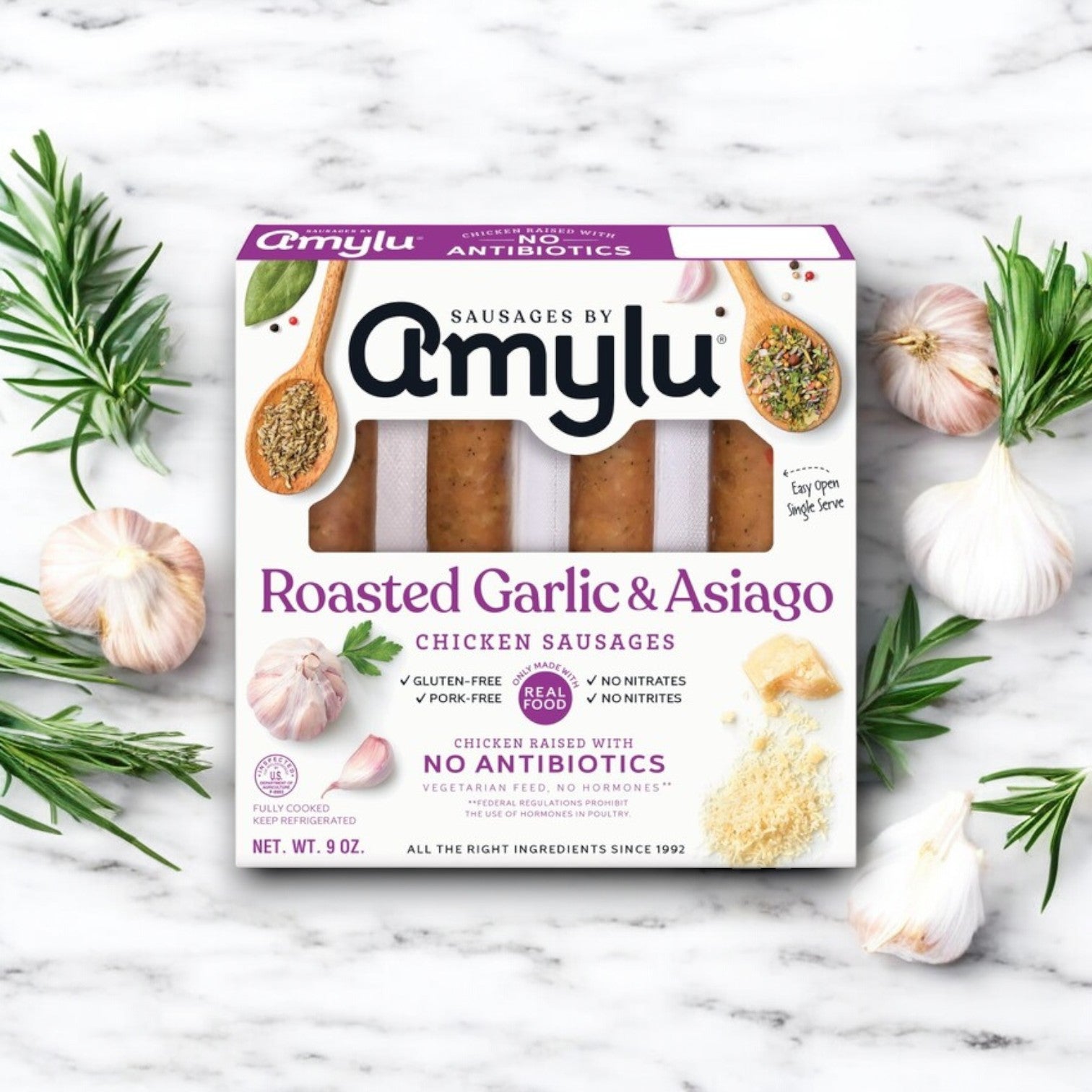 A package of Amylu Roasted Garlic & Asiago Chicken Sausage - 4 CT (9 oz) sits on a marble surface with rosemary sprigs and garlic cloves, highlighting its gluten-free and antibiotic-free qualities.
