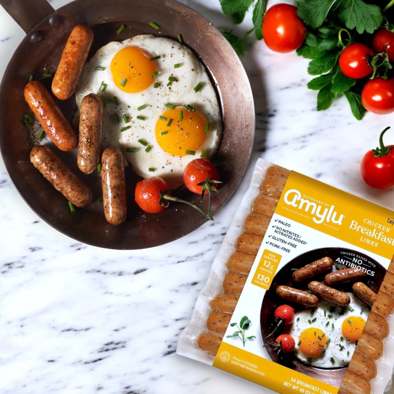 A frying pan with fried eggs, sausages, and tomatoes sits next to an Amylu Chicken Breakfast Sausage Links – 7.5 lbs package on a marble surface, surrounded by cherry tomatoes and fresh herbs. Enjoy a delicious, gluten-free start to your day!.