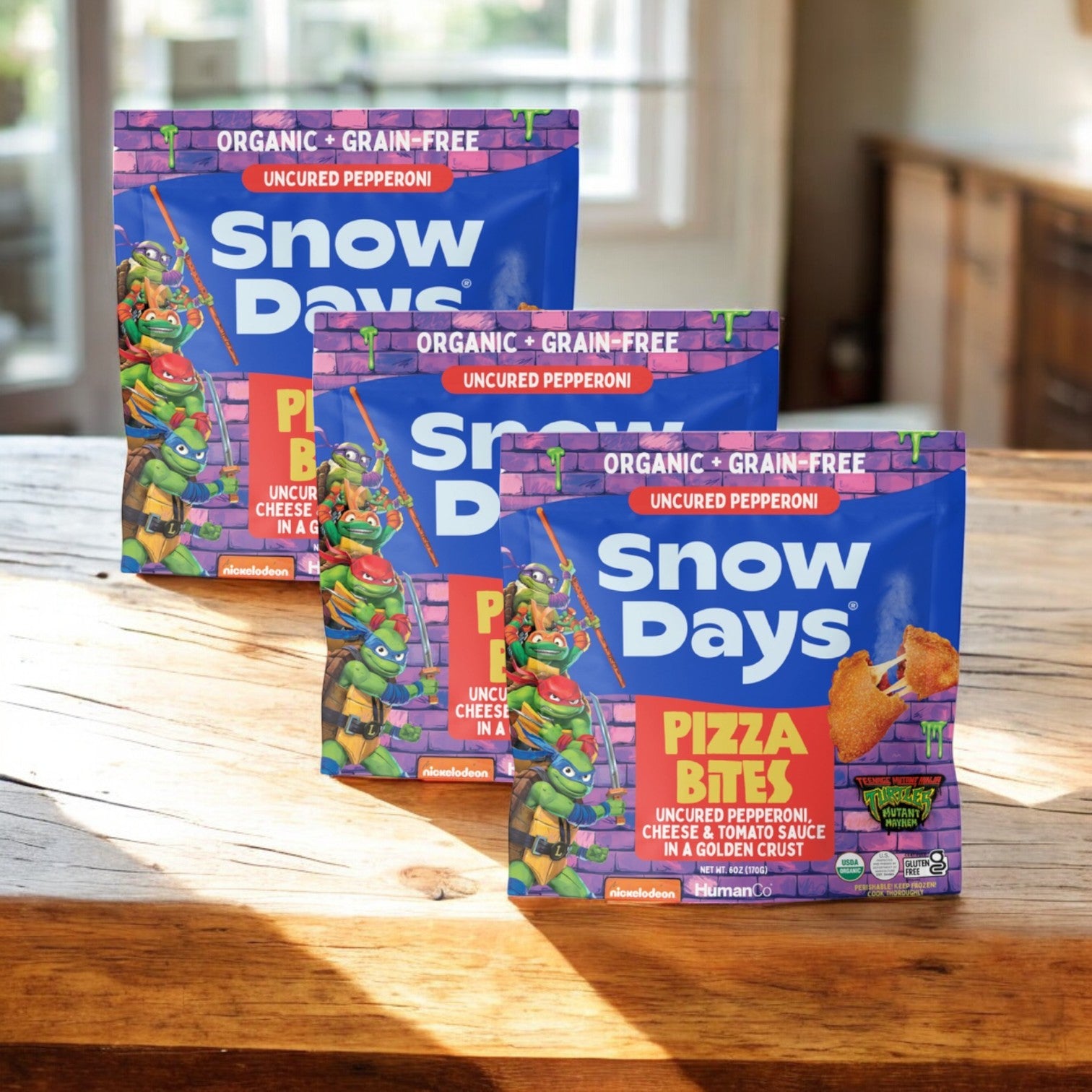 Three bags of Snow Days Uncured Pepperoni Pizza Bites, gluten free and grain free (6 oz. each), are displayed on a wooden surface with a blurred background.