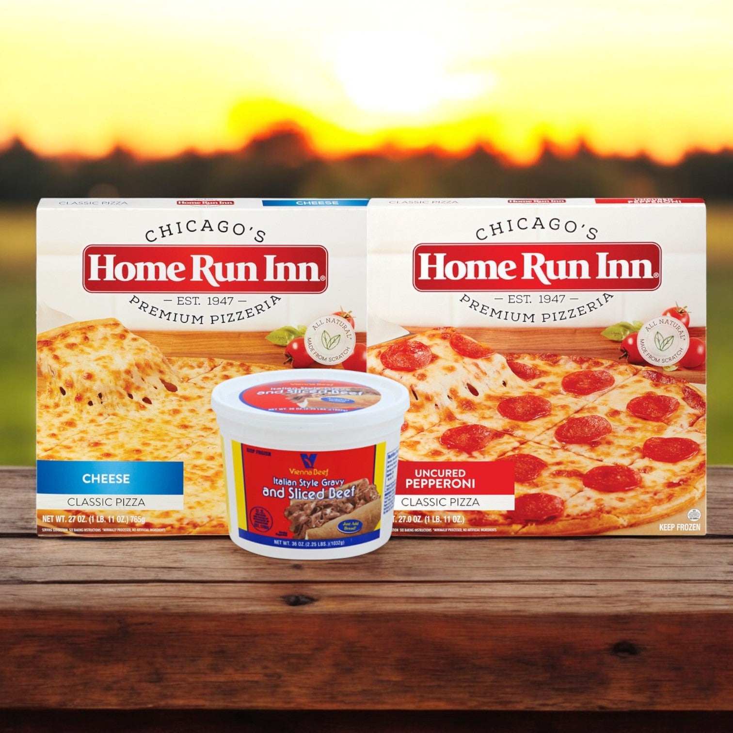 The Home Run Inn and Vienna Beef Chicago-Style Favorites Combo by Easy Lunches features two pizza boxes, cheese and uncured pepperoni, with a tub of sliced Italian beef on a wooden surface, set against a warm sunset.