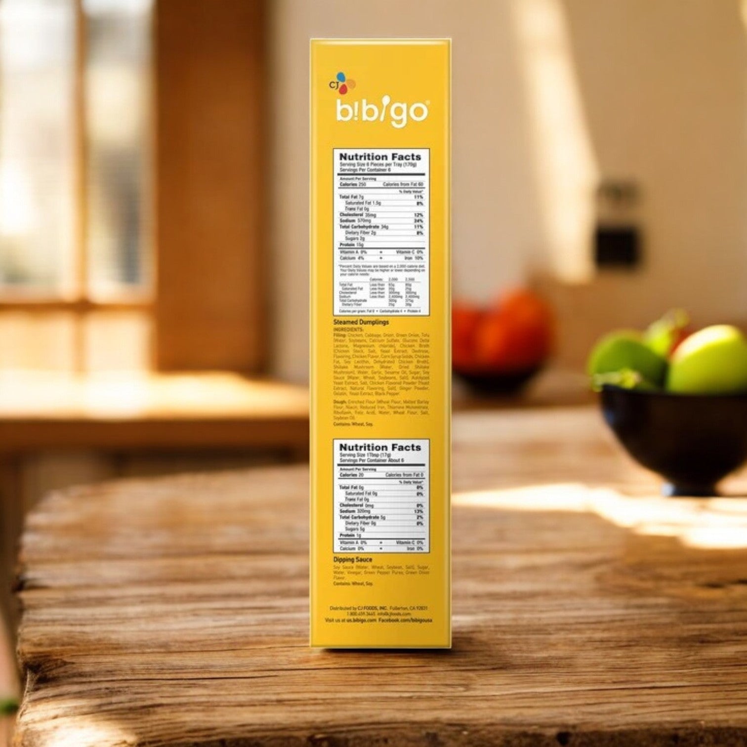 A 39.6 oz box of CJ Bibigo Steamed Dumplings, Chicken & Vegetable (18-count), stands upright on a wooden table with fruit blurred in the background, showing its nutrition facts on the side.