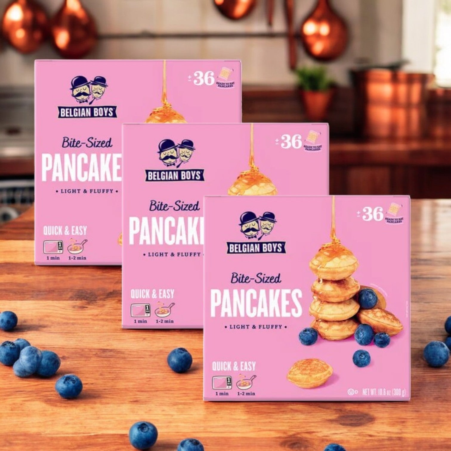 Three packs of Belgian Boys Refrigerated Bite-Sized Pancakes (3 count) are shown on a wooden surface with scattered blueberries and a kitchen background.