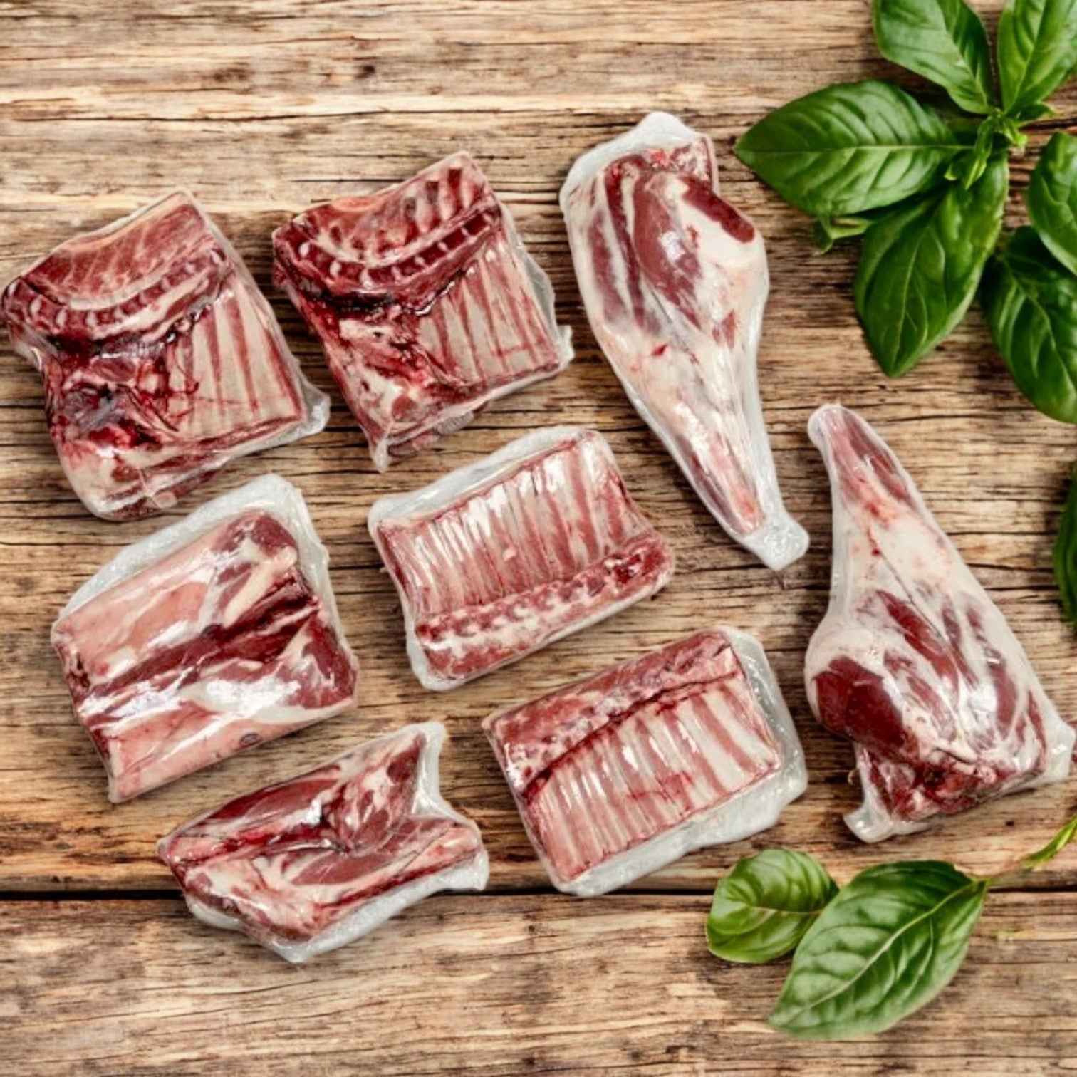 Several vacuum-sealed cuts of New Zealand Spring Lamb Halal Lamb Carcass, 8-Way Cut (28 lb avg wt), are arranged on a wooden surface beside fresh basil leaves, highlighting the freshness and quality of this premium product.