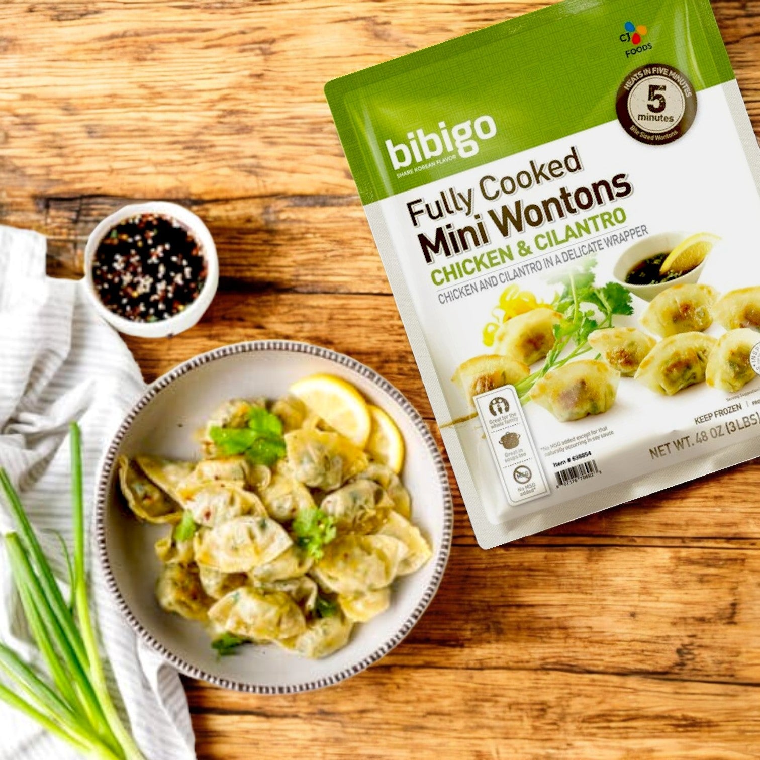 A 3-lb bag of Bibigo Mini Wontons – Chicken & Cilantro is set beside a bowl of Korean-style wontons with herbs and lemon slices, accompanied by green onions and a side dish.