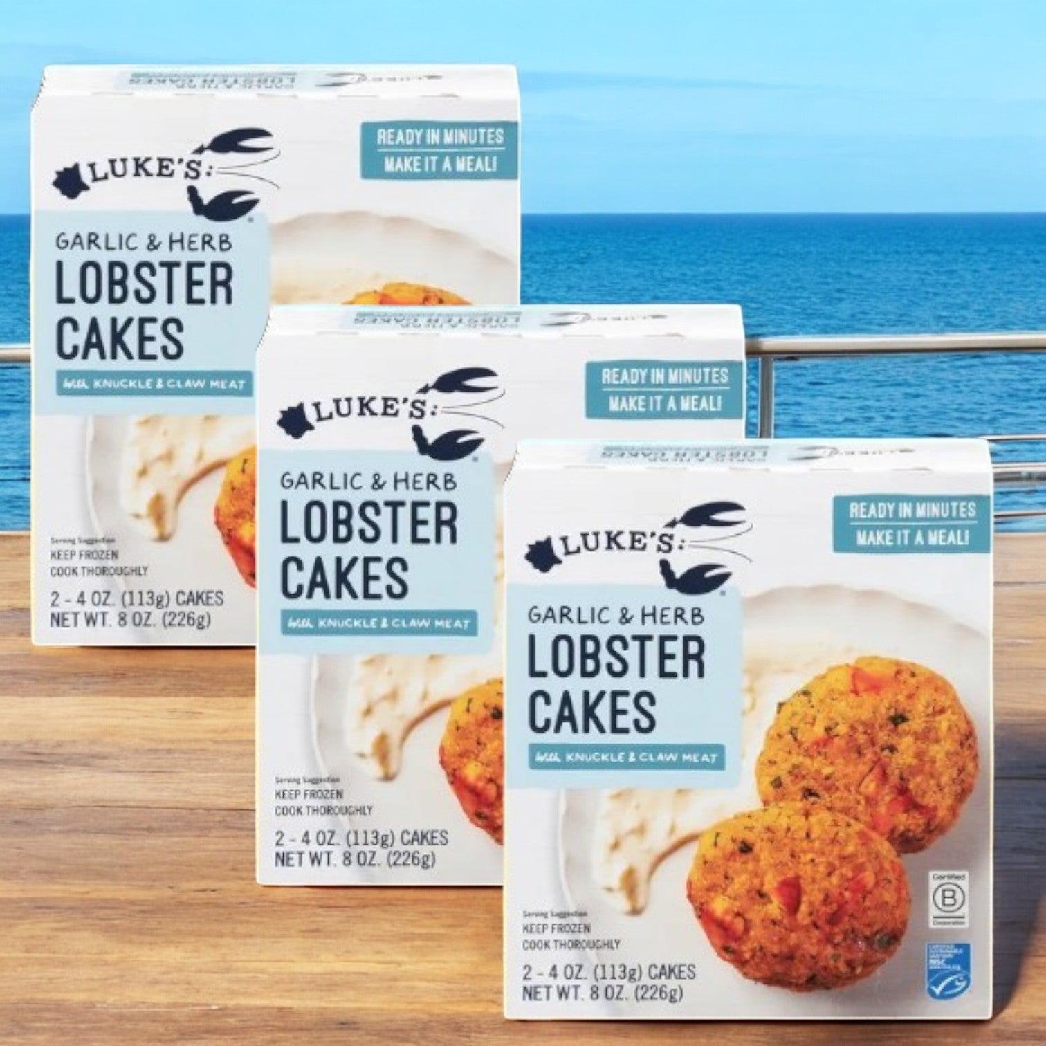 Three boxes of Luke's Organic Lobster Cakes, Garlic & Herb-3 Count are arranged on a table by the ocean, promising a delicious seafood experience.