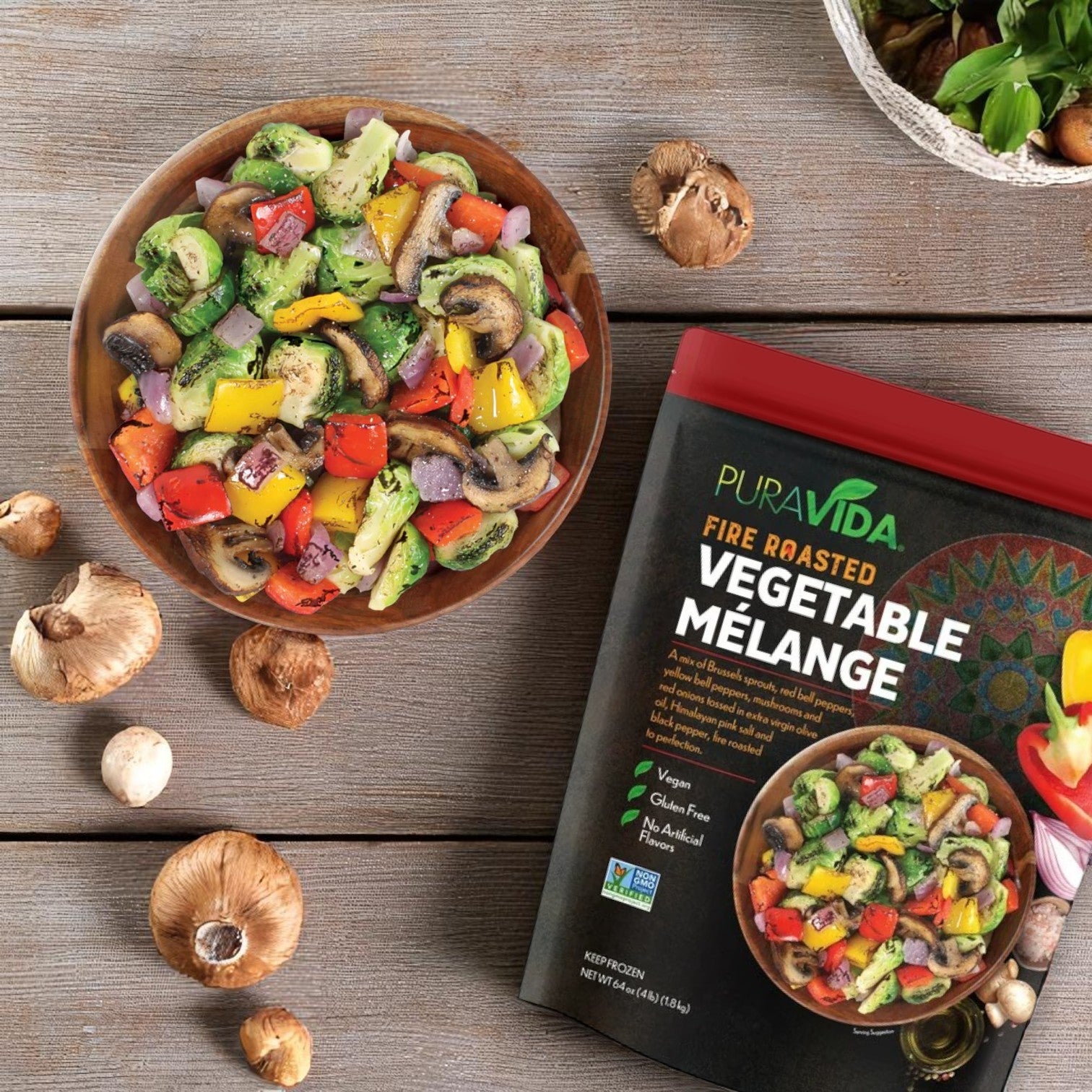 A bowl of mixed fire-roasted vegetables sits beside a "Pura Vidaa Fire-Roasted Vegetable Mélange – 3 Pack (64 oz Each)," showcasing this frozen blend as a healthy side dish on a wooden surface with scattered mushrooms.