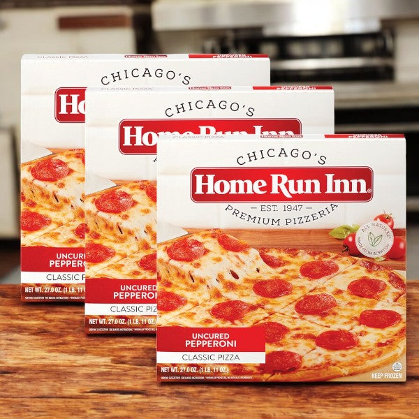 Three boxes of Home Run Inn Classic Pizza, Uncured Pepperoni, 27 oz. (3 total pizzas) are displayed on a kitchen counter with their signature homemade-style crust and a stove in the background.