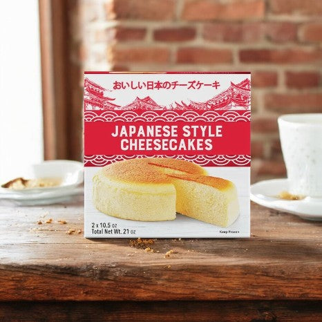 A Kirkland Signature Delcato Japanese Style Cheesecake box (2-count) sits on a wooden table, featuring images of fluffy cheesecakes, with teacups and a brick wall in the background.