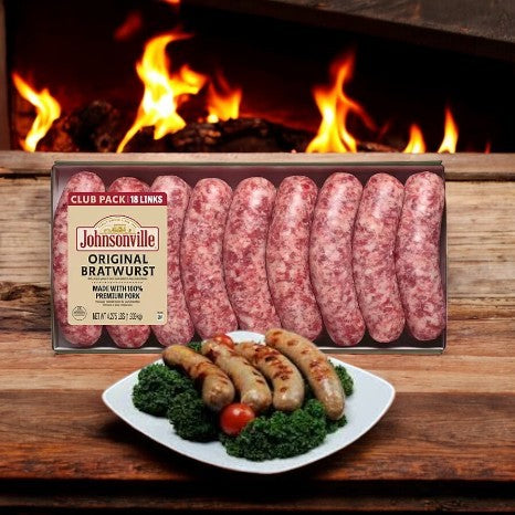 A case of Johnsonville Brats Original Pork Bratwurst Links (18 count) is shown behind a plate of cooked bratwursts with kale and cherry tomatoes, emphasizing their savory flavor with a cozy fireplace in the background.