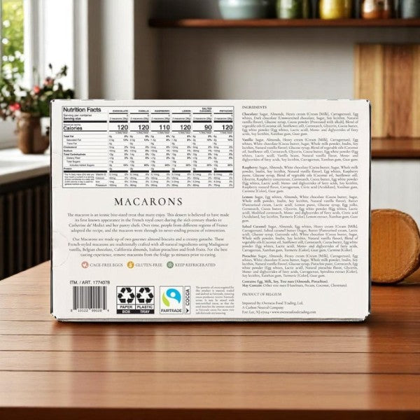 A box of Le Chic Patissier Macarons Variety, 36 Count – 3 Packs, is on a wooden countertop showing its nutrition facts, all-natural ingredient list, description, and certification symbols; kitchen shelves are visible behind.