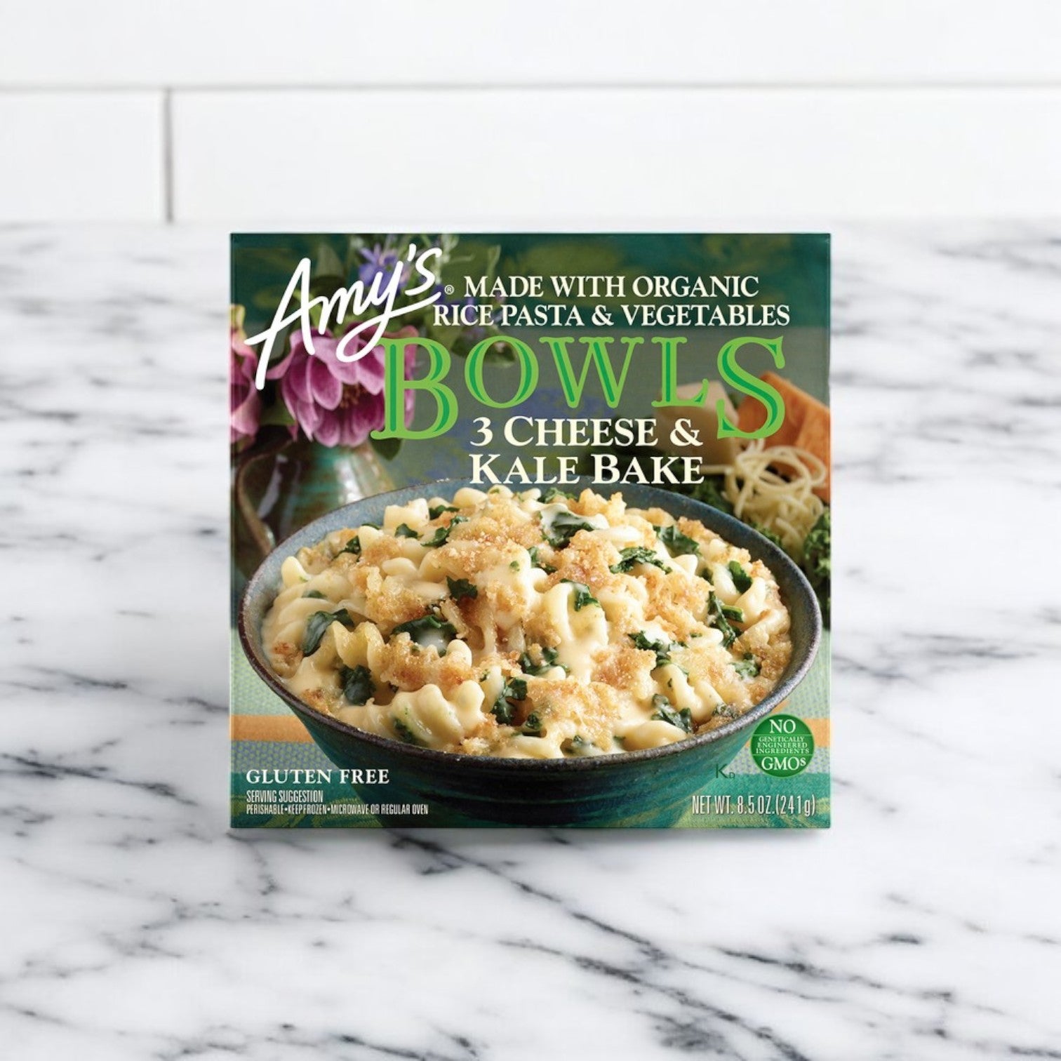 An 8.5 oz. box of Amy’s Kitchen Gluten Free Three Cheese Kale Bowl is displayed on a white marble surface, featuring organic ingredients and presenting this Amy's vegetarian meal as a tasty gluten-free option.