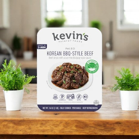 Kevin's Paleo Korean BBQ-Style Beef (32oz, 1 Count) by Kevin's is shown on a wooden table between two green potted plants in a kitchen, emphasizing its Asian-inspired flavors.